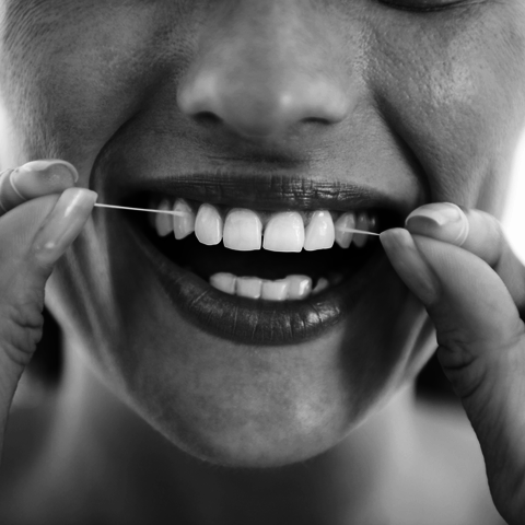 A close-up black-and-white photo of a person flossing their teeth, with a big smile showing clean, well-aligned teeth.