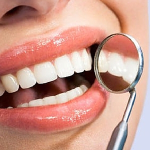 a person's mouth, showing clean, white teeth and healthy gums. A dental mirror is held up to reflect the teeth, highlighting their alignment and cleanliness. The overall impression is one of good oral hygiene and professional dental care.