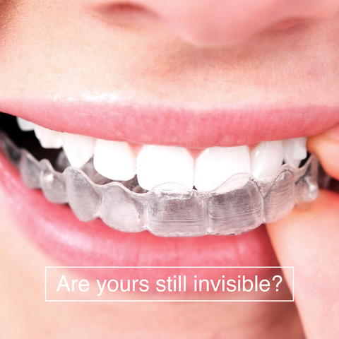 a close-up of a person's mouth with a clear dental aligner over their teeth. The aligner is transparent, fitting snugly over the teeth. Below the image, there is text that reads, "Are yours still invisible?"