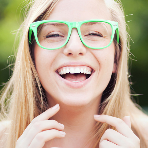 A joyful young woman with long blonde hair and bright green glasses smiles widely, showcasing her perfectly aligned teeth.