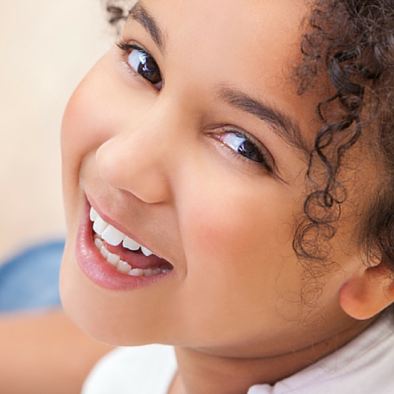 A close-up image of a smiling young girl with curly hair. She has bright, expressive eyes and a radiant smile, showcasing her clean, white teeth.