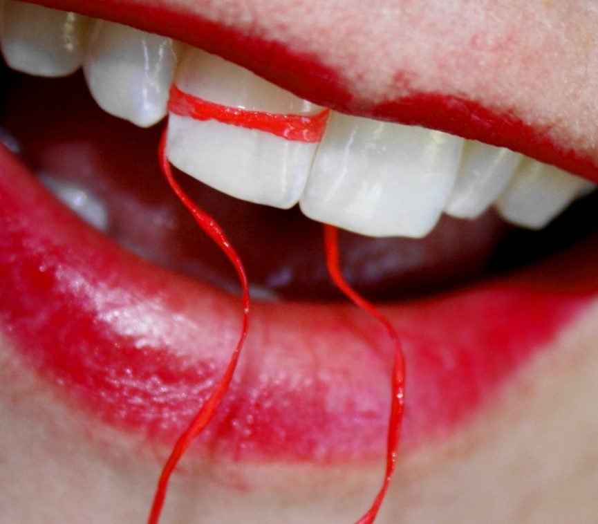a person's mouth with bright red lipstick. A piece of dental floss is shown wrapped around their front teeth, highlighting the importance of flossing. The teeth are clean and white, and the contrast between the red floss and lipstick against the white teeth.