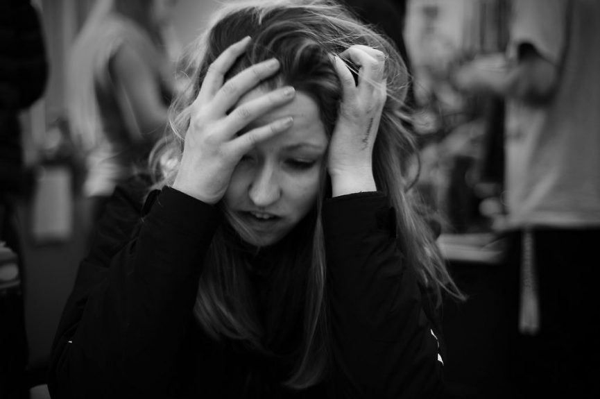 black and white image captures a woman in a moment of distress. She is sitting with her head in her hands, fingers tangled in her hair, and an expression of frustration or despair on her face. The background is blurred, focusing the viewer's attention on her emotional state.