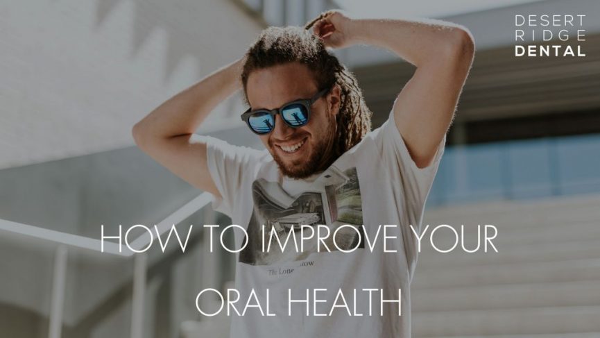 a man with dreadlocks and sunglasses, smiling broadly. He is wearing a white t-shirt with a graphic print and is standing outdoors in a sunny setting. The text overlay on the image reads "HOW TO IMPROVE YOUR ORAL HEALTH" in white, uppercase letters. The logo "DESERT RIDGE DENTAL" is placed in the upper right corner.