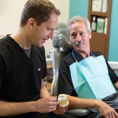 Two men are sitting at a desk in a dental office, discussing overdentures. On the desk are dental models, including one showing an overdenture. In the background, a monitor displays images of a patient's mouth before and after treatment, highlighting the dental work involved with overdentures.