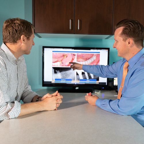 Dr. Rogers is wearing a light blue shirt and orange tie is seated across from a patient who is wearing a checkered shirt. Dr. Rogers is pointing to a monitor that displays "before and after" images of dental procedures, along with X-ray images. The patient is listening attentively, with hands clasped on the table. 