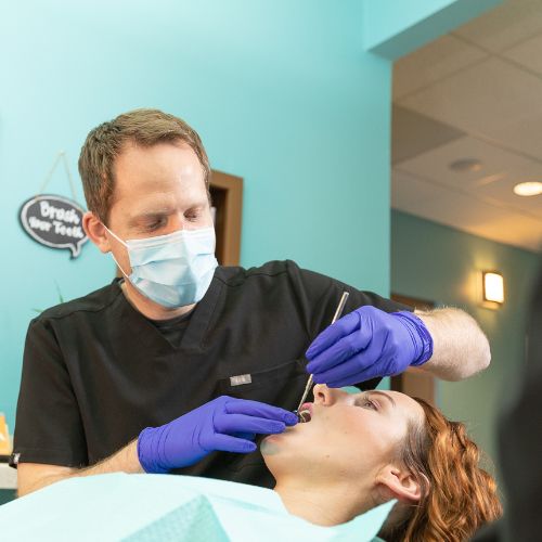 Dr. Mink, wearing a face mask, black scrubs, and blue gloves, is carefully examining a patient's teeth. The patient is lying back in the dental chair with a dental bib on, while the dentist uses dental tools to check her mouth. 