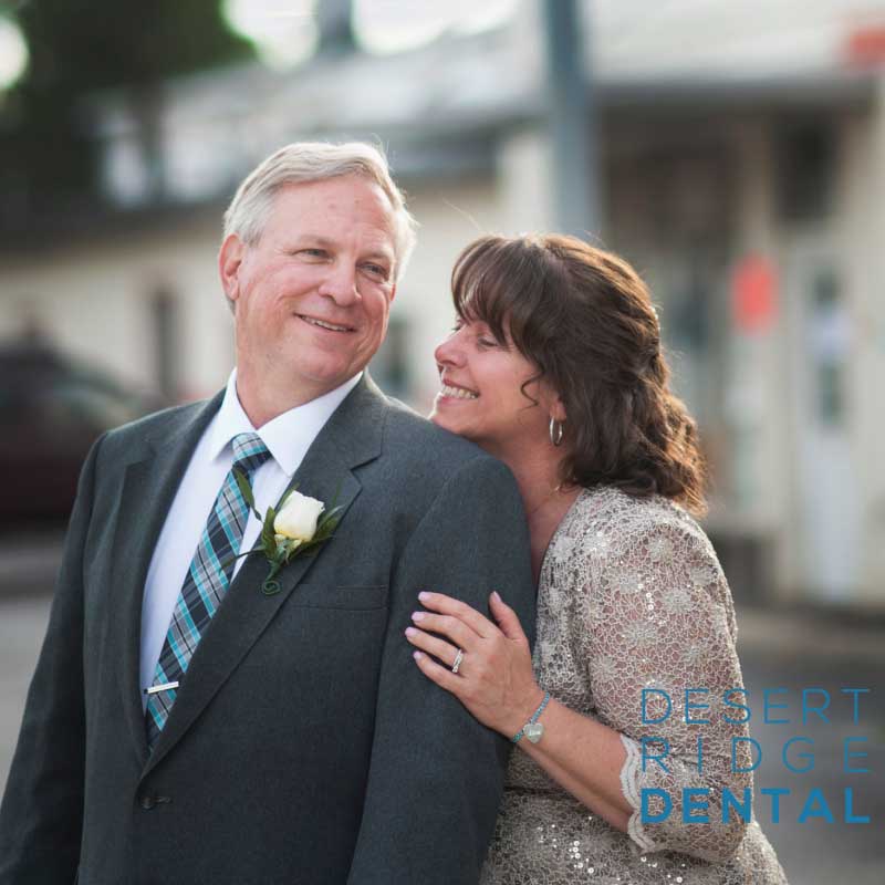 an older couple standing close together, smiling and looking at each other affectionately. The man is wearing a dark suit with a white shirt and a striped tie, and he has a white rose boutonniere pinned to his lapel. The woman, with shoulder-length brown hair, is dressed in a light-colored lace dress. She has her hand on the man's shoulder, and they both appear happy and content.
