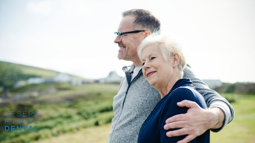 An older couple standing outdoors in a scenic, grassy area. The man, with short hair and wearing glasses, has his arm around the woman, who has short blonde hair. They are both dressed in casual, warm clothing. The woman is leaning her head on the man's shoulder, and they both appear content and happy. In the background, there are rolling hills and a few buildings, suggesting a rural or countryside setting.