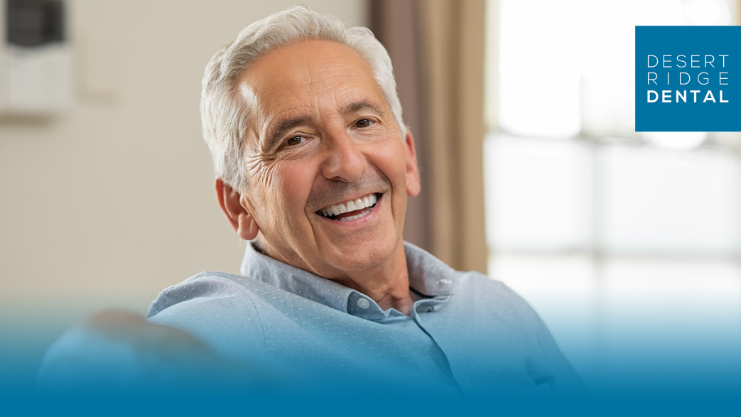 a smiling older man with gray hair sitting comfortably indoors. He is wearing a light blue shirt and looking directly at the camera, appearing happy and relaxed. The background is softly blurred, with a window and curtain visible. In the top right corner, there is the Desert Ridge Dental logo