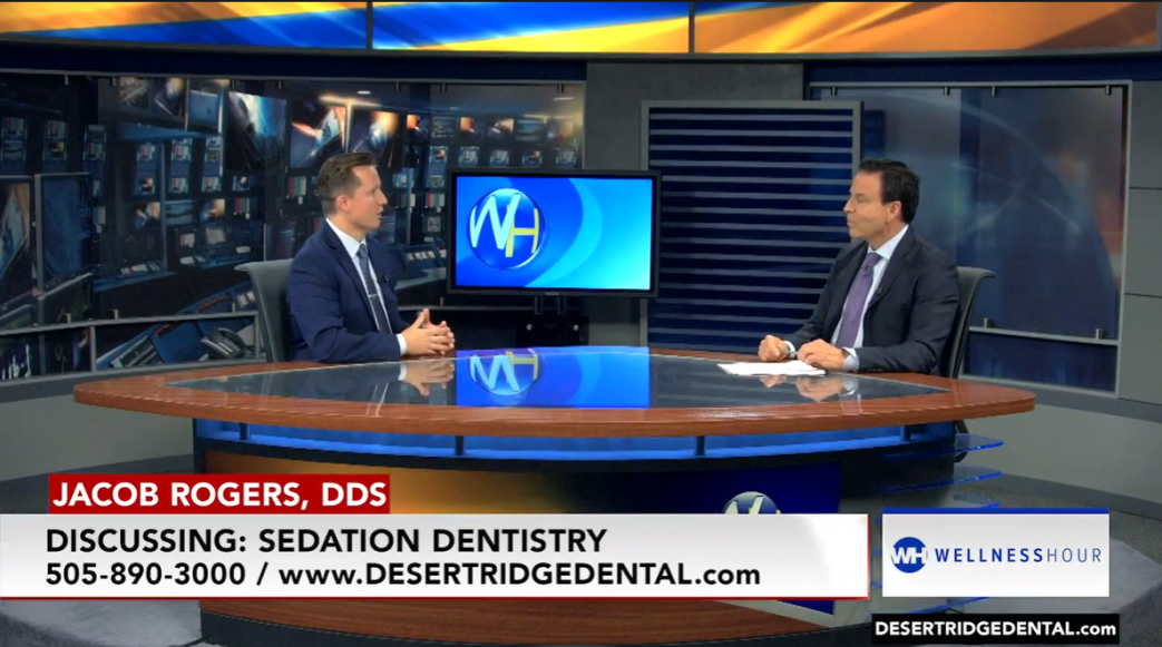 a television interview set where Jacob Rogers, DDS, is discussing sedation dentistry. Dr. Rogers is seated on the left side of a round table, wearing a suit and tie, and talking to the host, who is seated on the right, also dressed in a suit and tie. Behind them, a monitor displays the logo for "Wellness Hour." The bottom of the image has text that reads, "Jacob Rogers, DDS, Discussing: Sedation Dentistry, 505-890-3000 / www.DESERTRIDGEDENTAL.com."